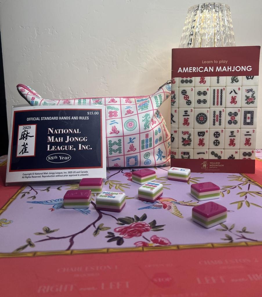 Mahjong tiles, National Mahjong League card, and American Mahjong beginner book arranged on a colorful Mahjong table.